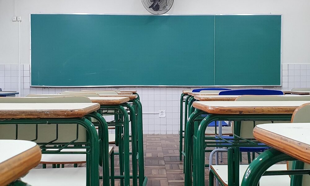 Vista de trás de uma sala de aula, com a lousa de frente para quem olha a imagem e um vão entre as carteiras, alinhadas lado a lado